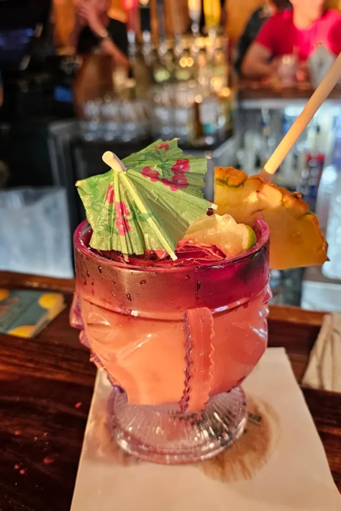 A vibrant tropical cocktail served in a unique glass with a sculpted face design, garnished with a green paper umbrella, a pineapple slice, and a lime wedge. A straw is placed in the drink, and the background features a blurred bar setting.