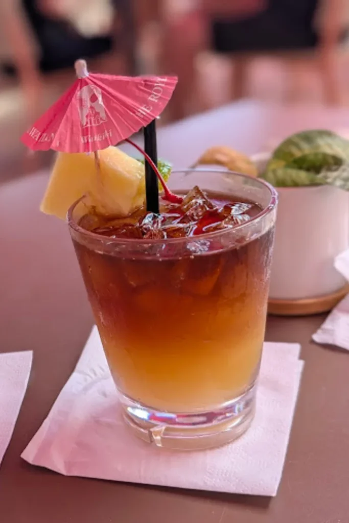 A classic Mai Tai cocktail in a short glass, featuring a dark rum float over golden liquid, garnished with a pink paper umbrella, a pineapple wedge, and a cherry. The drink is placed on a white napkin with a blurred indoor setting in the background.