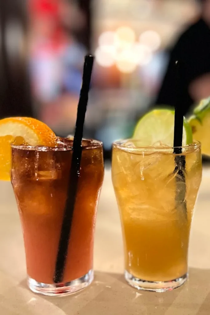 Two tropical cocktails side by side on a bar counter. The left drink is a dark rum-based beverage garnished with an orange slice, while the right drink is a lighter-colored cocktail with a lime garnish. Both have black straws, and the background is softly blurred with bar lights.