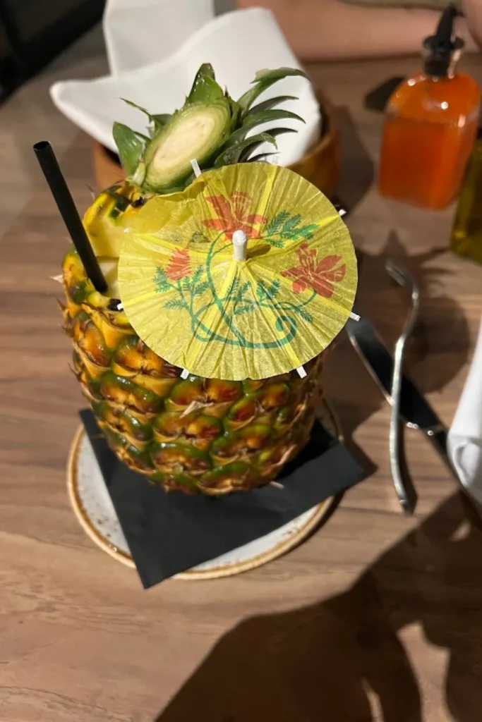 A tropical cocktail creatively served in a hollowed-out pineapple, topped with a decorative yellow paper umbrella featuring floral patterns. A black straw is inserted into the drink, and the setting includes a rustic wooden table with silverware and condiment bottles in the background.